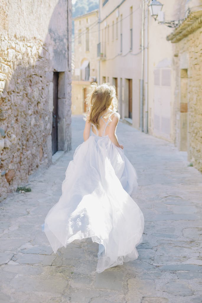 A woman in a dreamy, flowing dress runs down a narrow, sunlit old town street.